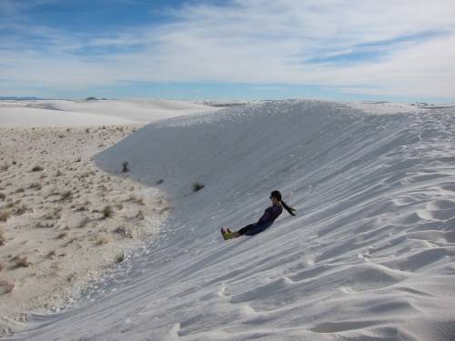 A girl slides down a white sand dune under a clear blue sky, surrounded by a vast desert landscape.