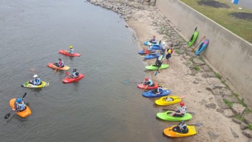 A group of kayakers in colorful kayaks gather by the shore, with some on the water and others on land.