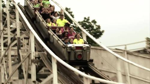 A group of excited riders on a roller coaster, descending a steep track with smiles and hands raised.