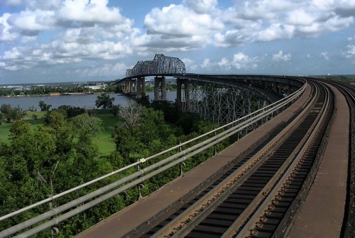 A railway bridge curves over a river, surrounded by greenery and under a partly cloudy sky.