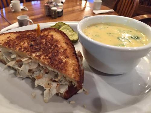 A toasted chicken sandwich with a pickle and a bowl of creamy soup on a white plate.