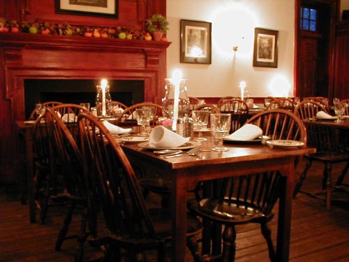Cozy dining room with wooden tables, chairs, candles, and decorative fruit on a mantel. Warm, inviting atmosphere.