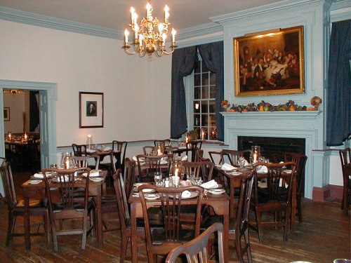 Cozy dining room with wooden tables, chairs, a chandelier, and a fireplace, featuring warm lighting and decorative artwork.