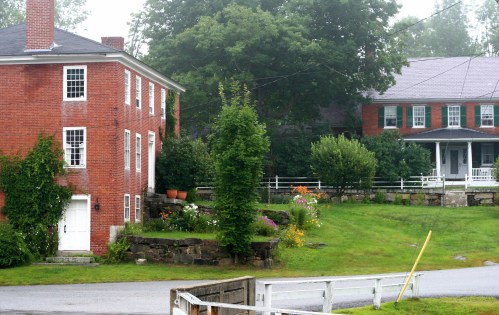 A foggy scene featuring two brick houses surrounded by greenery and colorful flowers in a rural setting.