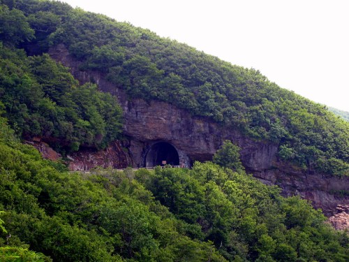 A tunnel entrance carved into a rocky hillside, surrounded by lush green trees and vegetation.