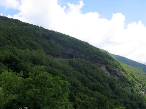 Lush green mountainside with a cave entrance, under a partly cloudy blue sky.