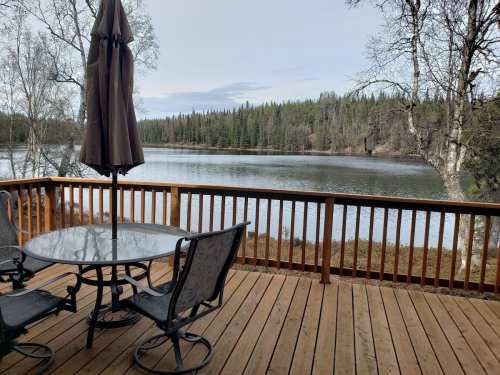 A wooden deck with a table and chairs overlooks a calm lake surrounded by trees and greenery.