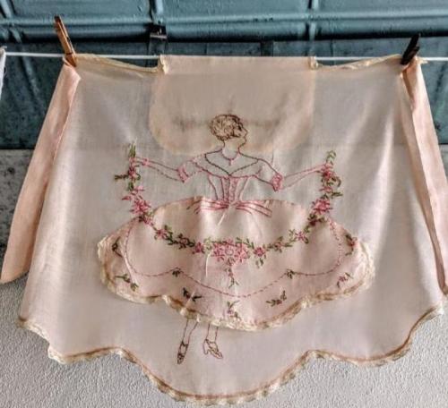 A vintage apron featuring an embroidered ballerina holding a floral garland, with a scalloped hem and soft pastel colors.