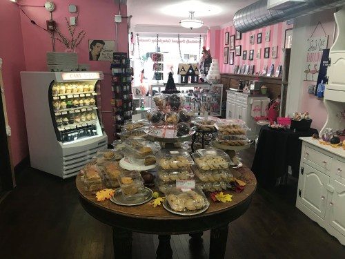 A cozy bakery with a pink interior, featuring a display of assorted baked goods on a table and a fridge with cakes.
