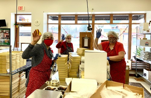 Three women in aprons and masks wave from a candy shop, surrounded by boxes and candy-making supplies.