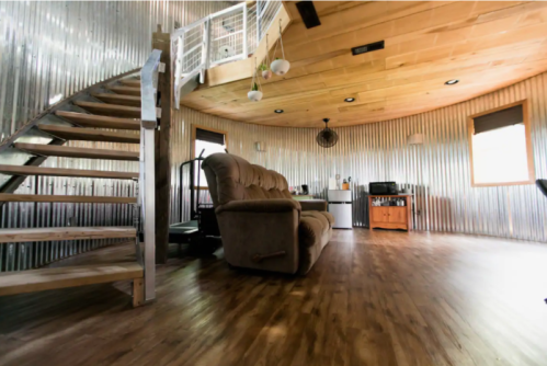 A cozy interior with a spiral staircase, a comfortable couch, and a kitchenette in a round, metallic-walled room.