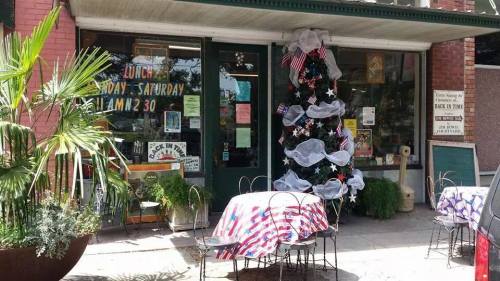 A quaint café entrance decorated with a patriotic Christmas tree and outdoor seating with red, white, and blue tablecloths.