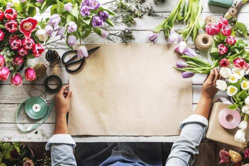 A person’s hands on a table surrounded by colorful flowers, scissors, and crafting materials, ready for floral arrangement.