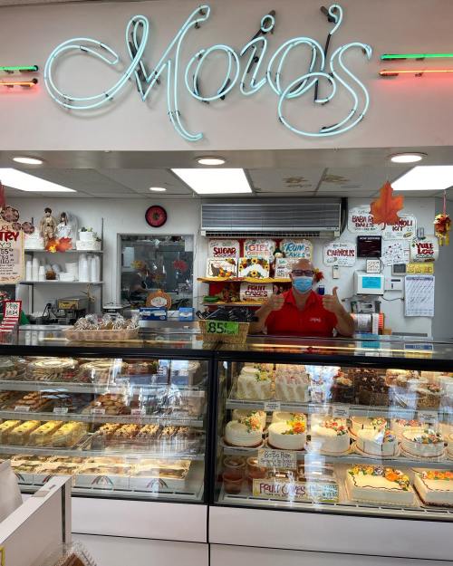 A bakery display filled with various pastries and cakes, with a smiling employee wearing a mask behind the counter.