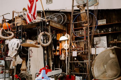 A cluttered antique shop filled with various vintage items, including bikes, hats, and a ladder against a wooden wall.