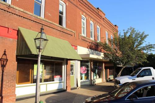 A brick building with green awnings, featuring storefronts and a lamp post, on a sunny day.
