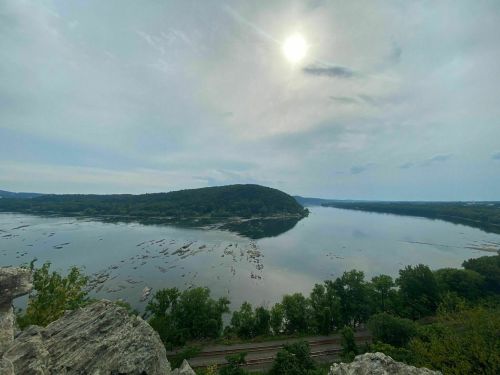 A serene river landscape under a cloudy sky, with lush greenery and distant hills reflected in the water.