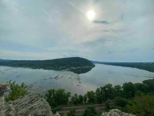 A serene river landscape under a cloudy sky, with lush greenery and distant hills reflected in the water.