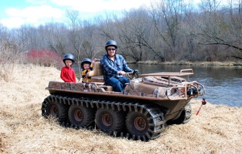 A man and two children sit in an all-terrain vehicle by a river, surrounded by bare trees and dry grass.