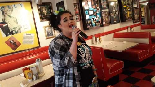 A singer performs with a microphone in a retro diner, surrounded by vintage photos and red booths.