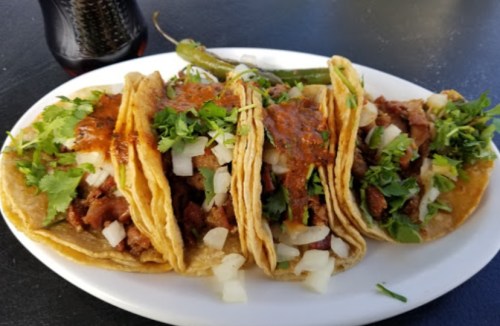 A plate of four tacos filled with meat, onions, and cilantro, topped with sauce, alongside a green pepper.