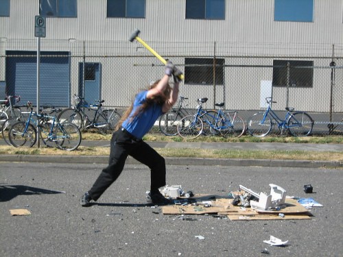 A person with long hair swings a sledgehammer at debris on the ground, with bicycles parked in the background.