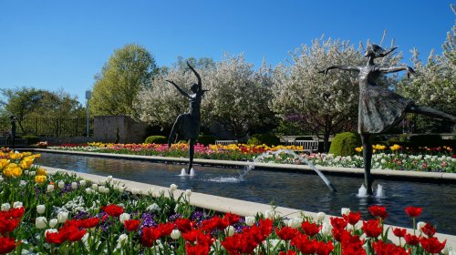 Two bronze statues of dancers beside a flower-filled garden with colorful tulips and a serene water feature.