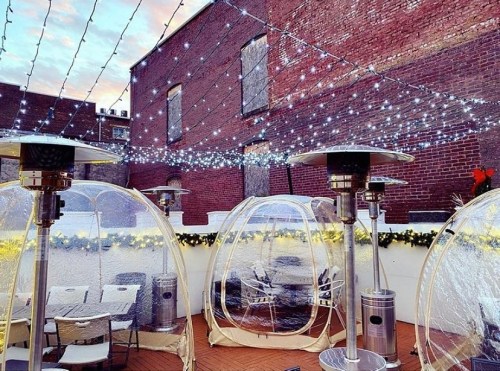 Outdoor dining area with clear igloo-like pods, string lights, and patio heaters, set against a brick wall.