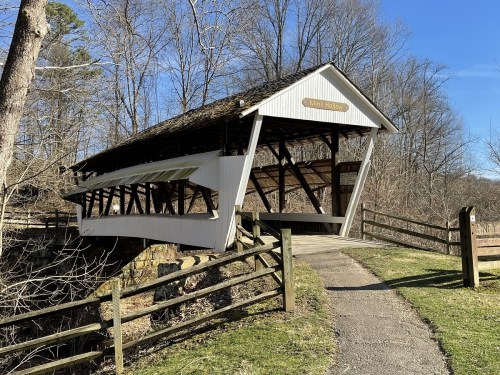 A covered bridge with a wooden structure, surrounded by trees and a grassy path leading to it. Clear blue sky above.