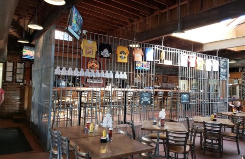 A rustic restaurant interior with a metal cage-like structure, decorated with T-shirts and a bar area in the background.