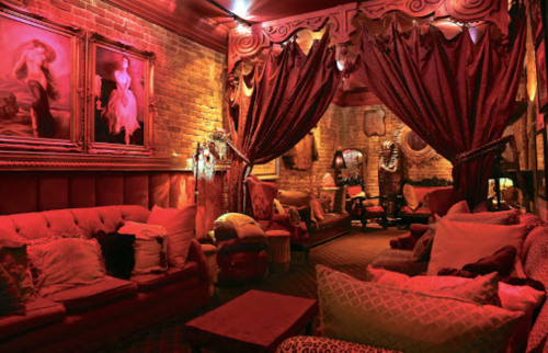 Cozy lounge with red lighting, plush seating, and ornate decor, featuring curtains and vintage artwork on the walls.