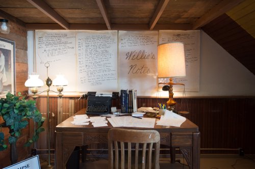 A cozy desk with a typewriter, scattered papers, and notes on the wall, illuminated by a warm lamp.