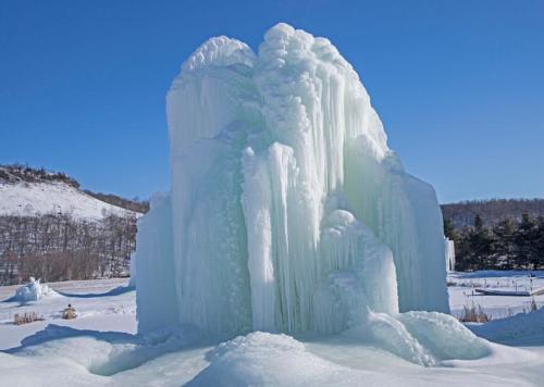 A towering ice formation glistens under a clear blue sky, surrounded by snow-covered hills and trees.