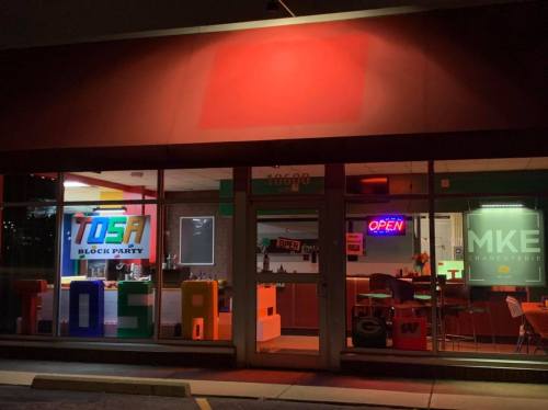 A brightly lit storefront at night featuring colorful signs for "TOSA BLOCK PARTY" and "MKE CHARCUTERIE."