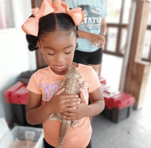A young girl in an orange shirt holds a lizard, smiling, with a person in the background.
