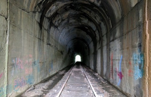 A dark, graffiti-covered tunnel with railway tracks leading to a bright light at the far end.