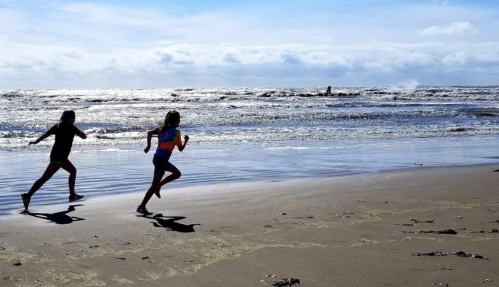 Two children run along a sandy beach, with waves crashing in the background under a bright blue sky.