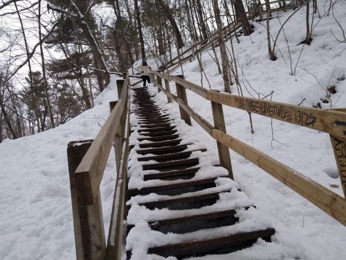 A snowy wooden staircase leads up a hill, surrounded by trees and a railing, with footprints visible in the snow.