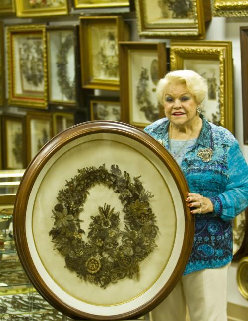 An older woman in a blue patterned jacket stands beside a large framed floral artwork, surrounded by more framed pictures.