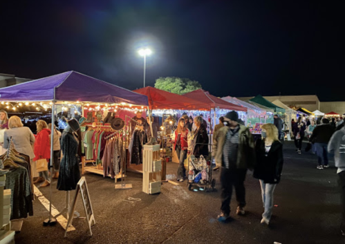 A night market scene with colorful tents, shoppers browsing clothing and goods under string lights.