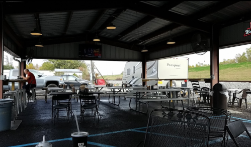 An outdoor dining area with empty tables, chairs, and a truck in the background under a metal roof.