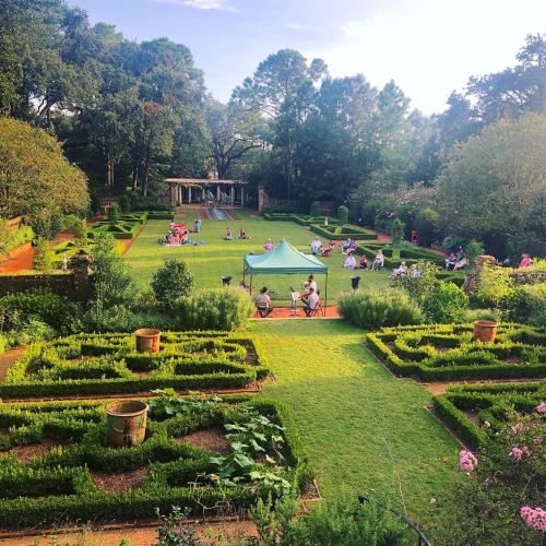 A lush garden with neatly arranged flower beds, people relaxing on the lawn, and a tent set up for an event.