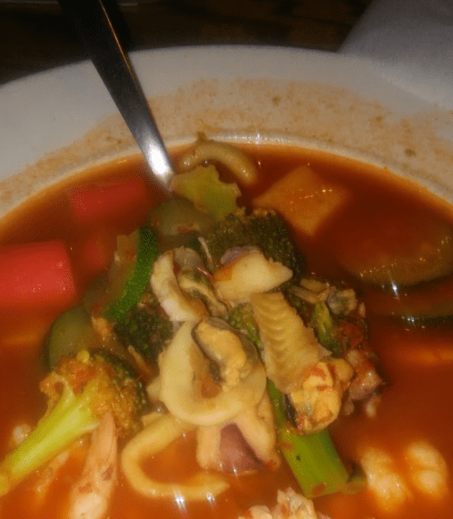A bowl of seafood soup with vegetables, featuring zucchini, broccoli, and various shellfish in a rich red broth.