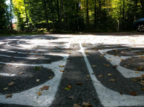A winding white maze painted on a gravel surface, surrounded by trees and scattered autumn leaves.