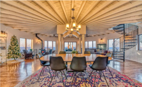 A spacious dining area with a round table, modern chairs, and a spiral staircase, featuring large windows and a Christmas tree.