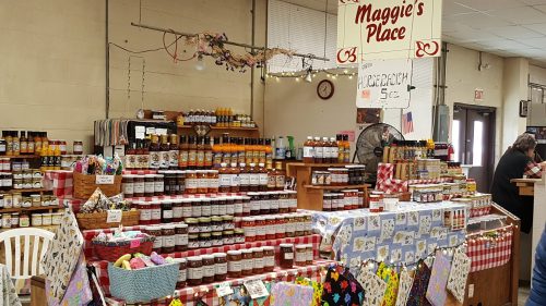 A vibrant market stall named "Maggie's Place" filled with jars of jams, sauces, and colorful handmade items.