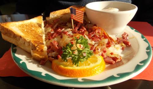 A plate of corned beef hash with toast, garnished with parsley, an orange slice, and a small bowl of sour cream.