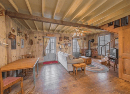Cozy interior of a rustic room with wooden walls, a sofa, dining table, and a staircase leading to an upper level.