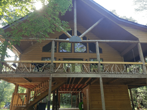 A large wooden cabin with a spacious deck, surrounded by trees and sunlight filtering through the leaves.