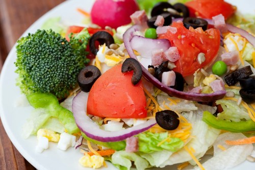 A colorful salad with lettuce, tomatoes, broccoli, olives, ham, onions, and hard-boiled eggs on a white plate.